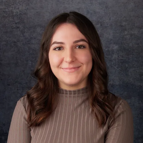 A smiling woman with medium-length brown hair wearing a high-necked, light-colored blouse poses confidently against a neutral gray background.