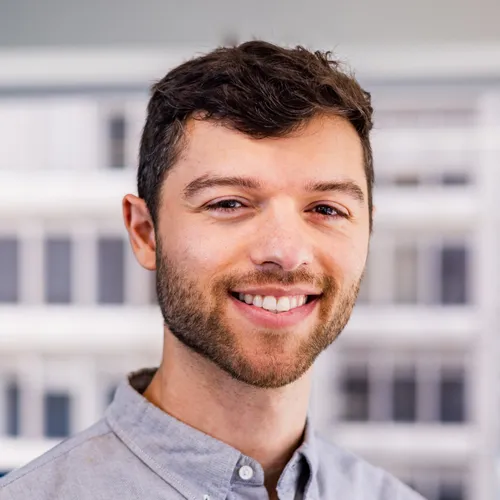 A smiling man with short dark hair and a light beard is wearing a light gray button-up shirt and posing outdoors against a blurred background of modern buildings.