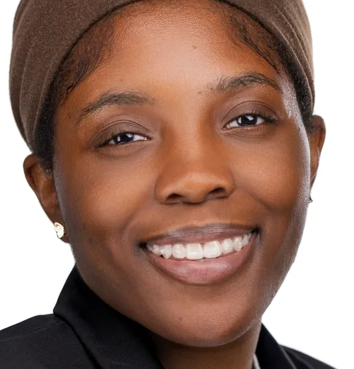 A smiling person with short, neatly styled hair tied back, wearing small earrings, and dressed in a dark top. The individual is smiling warmly with a bright and welcoming expression.