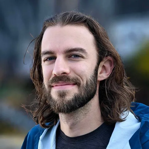 A person with long, wavy brown hair and a full beard smiling gently, wearing a dark shirt with a blue jacket partially visible.