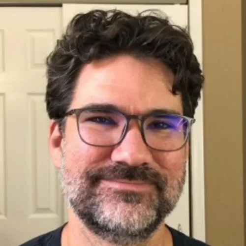 A person with short, wavy hair and a beard wearing glasses, looking directly at the camera with a friendly expression.