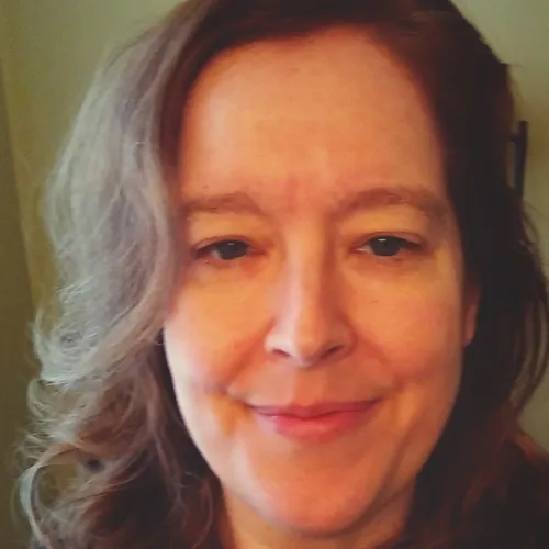 A person with shoulder-length brown hair smiling gently at the camera, showing a warm and friendly expression.